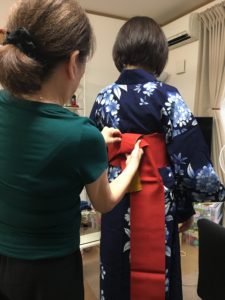 With their backs to us, an older woman ties the obi of a younger woman.