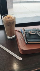 A clear glass of iced coffee sits next to a laptop case, black notebook, and white earbuds