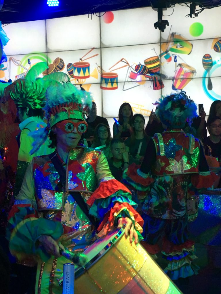 A performer with a large drum and colorful headdress. The audience watches from behind