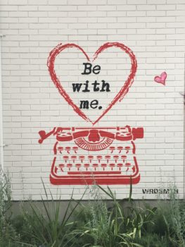 a white brick wall with a red spray painted typewriter. Above the typewriter is a heart that says "Be with me"