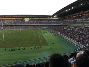 A large field with rugby players on the pitch. The sun is starting to set in the background
