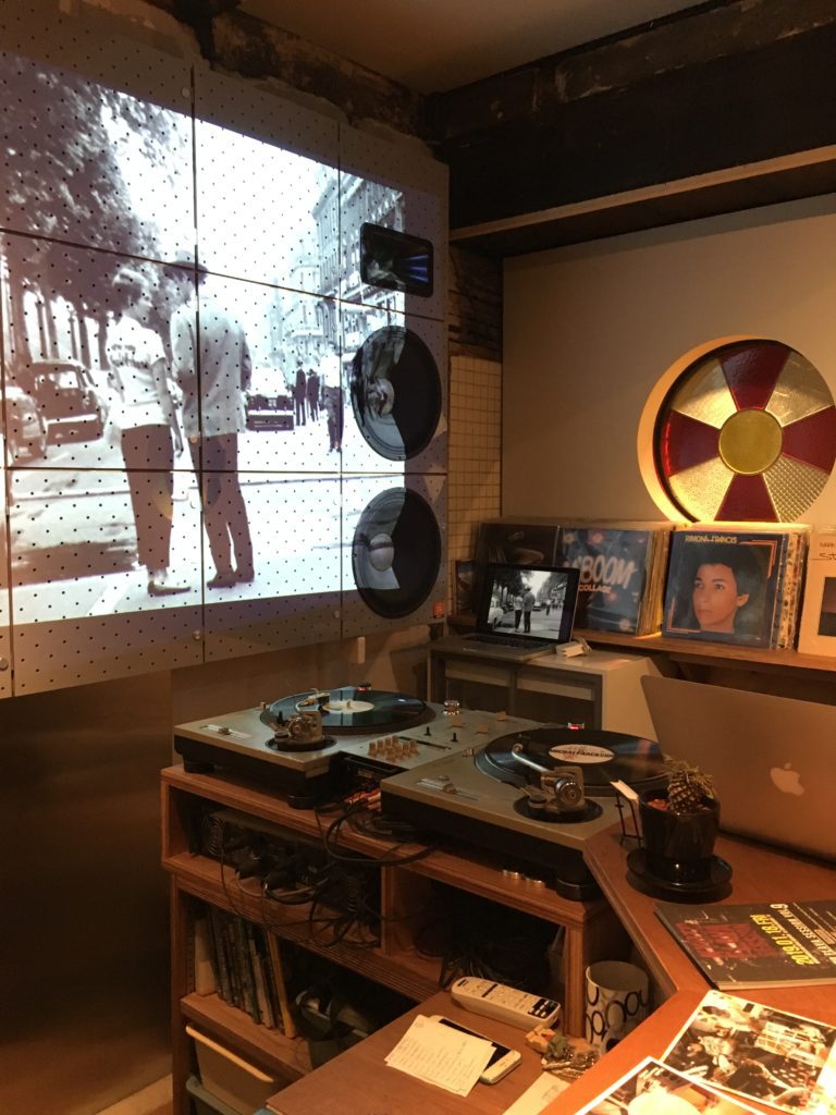 A bar with a black and white movie playing in the background. The end of the bar has a macbook and two turntables