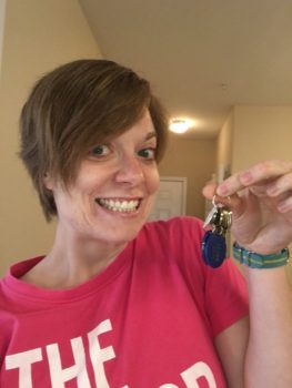 A woman with short hair, wearing a pink tshirt, holds up keys. She is smiling at the camera.