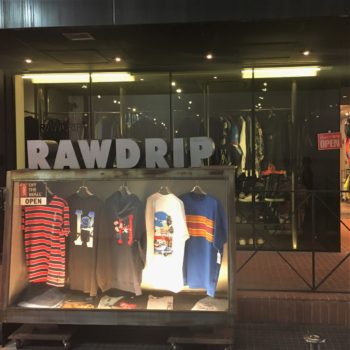 The exterior of Raw Drip thrift store features a lighted case outside with hanging t-shirts for sale